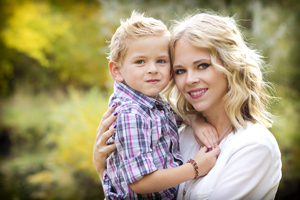 This is a picture with a younger lady with blonde hair and a white shirt holding a little boy in a black, white and red striped button up shirt. There is tear leaves in the background.