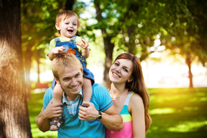 This is a picture of a couple and a toddler in a park. The lady is brown haired with a pink, yellow, blue and white tanktop, while the male has blonde hair and a blue shirt. The toddler is sitting on the males shoulders, he has light brown hair with a yellow shirt and overalls on.