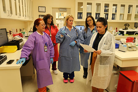 This is a picture of five lady doctors in a lab. There is two rolls on the far left side in the back is a lady with long brown hair in a orange coat, in the middle is a lady with blonde hair in a blue coat on the far right is a lady with glasses and a blue coat. in the front roll is a lady with red hair and glasses with a purple coat on the left, on the right is a lady with brown hair in a white coat reading a paper while everybody else is listening to her
